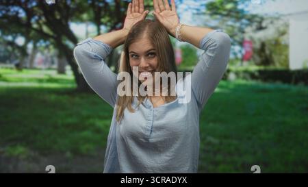 Blonde Frau hält Hände über dem Kopf und bildet Kaninchenohren, während sie im grünen Wald lächelt; spontane Freude. Stockfoto