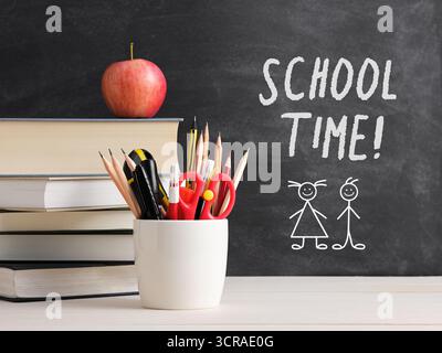 Schulzeitkonzept mit Apfel, Büchern und Schreibwaren vor einer Tafel mit der Schreibschulzeit und Kinderzeichnungen Stockfoto