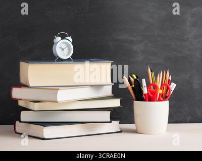 Schulbedarf einschließlich Wecker, Bücher und Bleistifte, die auf einem Schreibtisch vor einer leeren Tafel sitzen und Bildung und Lernen repräsentieren Stockfoto