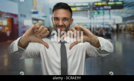 Junger Mann in einem weißen Hemd und einer Brille in einer urbanen Straßenlage, der mit einem Lächeln die Daumen nach unten drückt und im Freien eine ungezwungene Atmosphäre in der Stadt einfängt Stockfoto