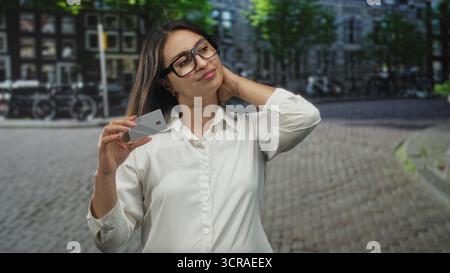 Frau, die die Kreditkarte in der Nähe der Kamera hält, während sie mit freier Hand auf einer kopfsteingepflasterten Straße in der Stadt Gesten macht, Brille trägt; Zahlungsfrustration. Stockfoto