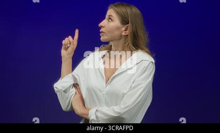 Die Frau zeigt den Finger im blauen Studio nach oben, trägt ein weißes Hemd mit gerollten Ärmeln und gefalteten Armen, blickt mit konzentriertem Ausdruck nach oben; Neugier. Stockfoto