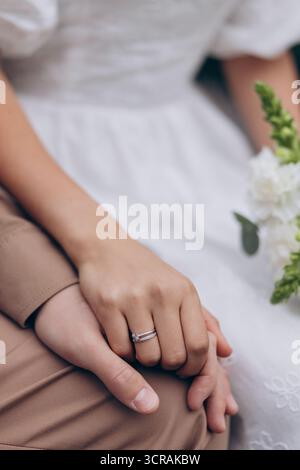 Nahaufnahme eines frisch verheirateten Paares, das zärtlich die Hände hält und die Details des Diamanten-Eherings und des weißen Brautkleides vor dem hervorhebt Stockfoto