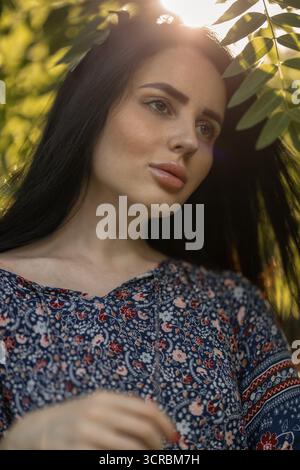 Die junge Frau steht anmutig zwischen üppig grünen Blättern in einem Blumenkleid. Das Sonnenlicht scheint durch das Laub und hebt ihre Gesichtszüge hervor, während sie nachdenklich in die Ferne blickt. Stockfoto