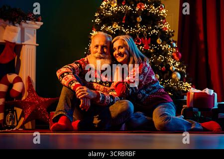 Ein liebevolles älteres Paar sitzt in der Nähe eines wunderschön dekorierten Weihnachtsbaums in einer gemütlichen Atmosphäre mit Geschenken und warmen Lichtern, die eine festliche Stimmung schaffen Stockfoto