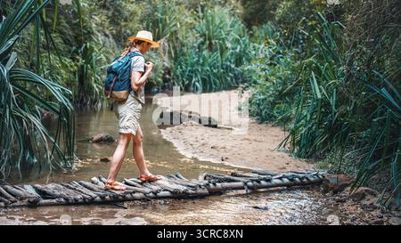 Junge Frau in beigefarbenem Hemd, Shorts und Strohhaw, Rucksack auf den Schultern, Wandern im Regenwald-Dschungel, Spaziergang über eine niedrige Holzbrücke am kleinen Bach in der Nähe Stockfoto