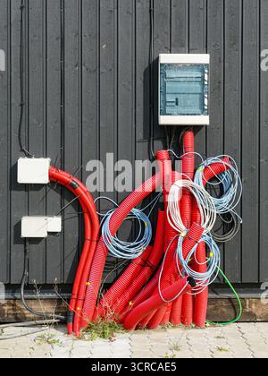 Elektrische Installation im Freien mit roten Rohrleitungen, Spiralkabeln und Sicherungskasten an einer schwarzen Holzwand Stockfoto