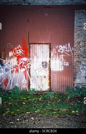 Große rote Metall-Lagerhaustüren, die nachts mit Graffiti bedeckt sind, mit direktem Blitz auf Analogfilm fotografiert, um Textur und urbanen Verfall zu betonen. Stockfoto