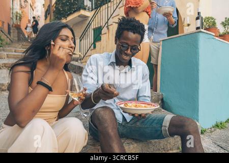 Ein glückliches junges Paar zwischen Rassen lacht, während es sich einen Teller frischer Pasta und Wein teilt und die lokale italienische Küche genießt, während es auf Stufen durin sitzt Stockfoto