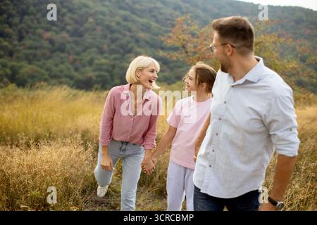 Mutter und Tochter lachen zusammen, während sie bei einem gemütlichen Spaziergang auf einem grasbewachsenen Feld die Hände halten. Ein Vater geht neben ihnen und lächelt ihn an Stockfoto
