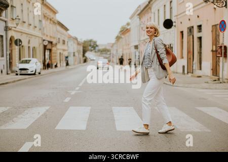 Eine Frau mit kurzen Haaren schlendert über einen Quersteg in einem ruhigen Stadtgebiet. Sie trägt legere Kleidung und trägt eine Handtasche, um einen angenehmen Tag zu genießen Stockfoto