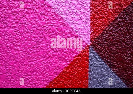 Mehrfarbige strukturierte Wand mit dreieckigen Mustern in leuchtenden Rosa-, Lila-, Rot- und Grautönen. Die Oberfläche weist eine raue, ungleichmäßige Aggregattextur auf Stockfoto