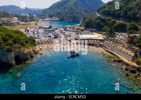 Drohnenblick auf den beliebten Strand Agios Spyridon in Paleokastritsa, Korfu. Eine geschäftige Sommerszene mit einem vollen Parkplatz, Touristen am Strand Stockfoto