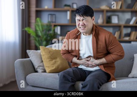 Mann, der seinen Bauch umklammert und auf einer Wohnzimmercouch grimmt, unter starken Magenschmerzen und Unwohlsein durch Verdauungsstörungen oder andere Magen-Darm-Erkrankungen zu Hause leidet Stockfoto