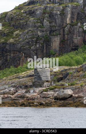 Old wooden coastal structure stands on rocky shore against steep cliffs with moss and greenery Stockfoto