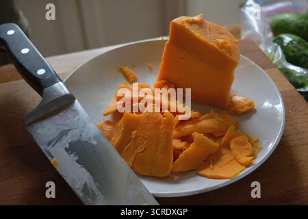 Scharfe Cheddar-Scheiben auf einer weißen Platte mit einem Küchenmesser – rustikale Szene zur Zubereitung von Speisen mit natürlichem Licht Stockfoto
