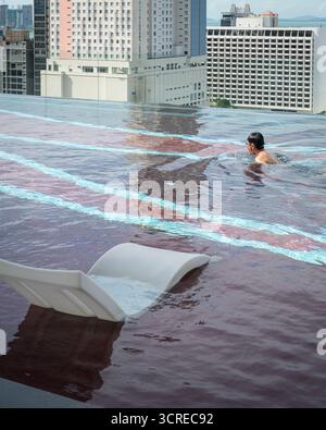 Mann schwimmt im Infinity-Pool auf dem Dach, mit Blick auf die Stadt im Hintergrund. Stockfoto