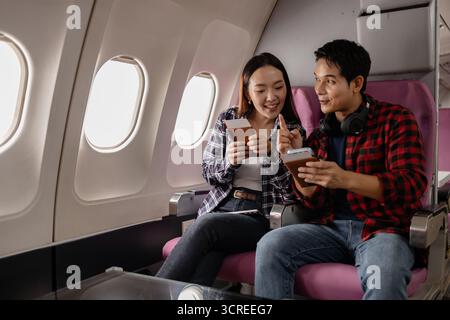 Ein junges asiatisches Paar, das im Flugzeug sitzt und lächelt, während er Pässe und ein Smartphone hält. Sie scheinen aufgeregt und bereit für ihre Reise zu sein, kleidet sich Stockfoto