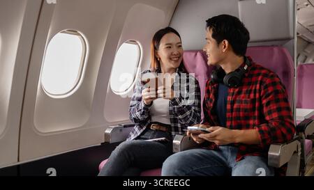 Ein junges asiatisches Paar, das im Flugzeug sitzt und lächelt, während er Pässe und ein Smartphone hält. Sie scheinen aufgeregt und bereit für ihre Reise zu sein, kleidet sich Stockfoto