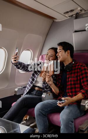Ein junges asiatisches Paar, das im Flugzeug sitzt und lächelt, während er Pässe und ein Smartphone hält. Sie scheinen aufgeregt und bereit für ihre Reise zu sein, kleidet sich Stockfoto