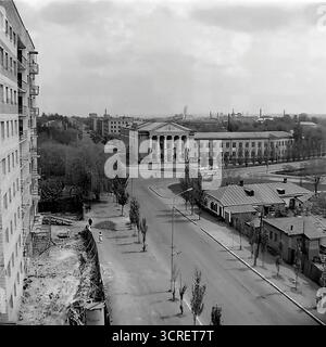 Ein Hochwinkel-Schwarzweiß-Archivfoto aus den 1980er Jahren, das einen Panoramablick auf Sloviansk in der ukrainischen SSR bietet. Der Mittelpunkt ist das monumentale Gebäude des Landwirtschaftskollegs, ein Paradebeispiel der neoklassischen Architektur des Stalinismus mit seinem großen Säulengang. Das Bild fängt die umliegende Stadtlandschaft der Svobody Street ein und zeigt einen Kontrast zwischen dem Grand College, funktionalen Wohnblöcken aus der Sowjetzeit und einer nahe gelegenen Baustelle. Die industrielle Skyline im Hintergrund stellt die Stadt fest in den Donbass und zeigt ihre einzigartige Mischung aus educ Stockfoto