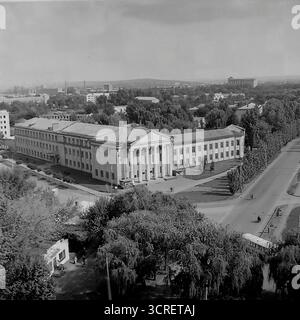 Ein Hochwinkel-Schwarzweiß-Archivfoto aus den 1980er Jahren, das einen Panoramablick auf Sloviansk in der ukrainischen SSR bietet. Der Mittelpunkt ist das monumentale Gebäude des Landwirtschaftskollegs, ein Paradebeispiel der neoklassischen Architektur des Stalinismus mit seinem großen Säulengang. Das Bild fängt die umliegende Stadtlandschaft der Svobody Street ein und zeigt einen Kontrast zwischen dem Grand College, funktionalen Wohnblöcken aus der Sowjetzeit und einer nahe gelegenen Baustelle. Die industrielle Skyline im Hintergrund stellt die Stadt fest in den Donbass und zeigt ihre einzigartige Mischung aus educ Stockfoto