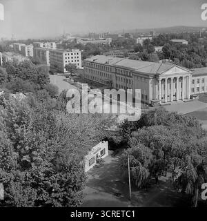 Ein Hochwinkel-Schwarzweiß-Archivfoto aus den 1980er Jahren, das einen Panoramablick auf Sloviansk in der ukrainischen SSR bietet. Der Mittelpunkt ist das monumentale Gebäude des Landwirtschaftskollegs, ein Paradebeispiel der neoklassischen Architektur des Stalinismus mit seinem großen Säulengang. Das Bild fängt die umliegende Stadtlandschaft der Svobody Street ein und zeigt einen Kontrast zwischen dem Grand College, funktionalen Wohnblöcken aus der Sowjetzeit und einer nahe gelegenen Baustelle. Die industrielle Skyline im Hintergrund stellt die Stadt fest in den Donbass und zeigt ihre einzigartige Mischung aus educ Stockfoto