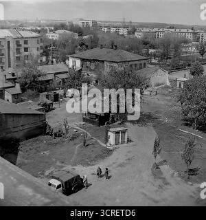 Ein Schwarzweiß-Archivfoto aus den 1980er Jahren, aufgenommen aus einem hohen Winkel in Sloviansk, Ukrainische SSR. Das Bild zeigt einen starken visuellen Kontrast zwischen zwei Epochen: Einem traditionellen, alten Innenhof mit heruntergekommenen Flachhäusern, Schuppen und unbefestigtem Boden und den modernen, mehrstöckigen sowjetischen Wohnblocks, die darüber ragen. Auf dem Hof befinden sich ein paar Bewohner und ein alter Nutzwagen, der eine vergangene Lebensweise darstellt, die von der neuen Stadtentwicklung überzogen wird. Diese Szene ist ein ergreifender Blick auf die städtische Transformation in einer Provinzstadt der Sowjetunion Stockfoto
