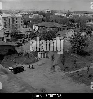 Ein Schwarzweiß-Archivfoto aus den 1980er Jahren, aufgenommen aus einem hohen Winkel in Sloviansk, Ukrainische SSR. Das Bild zeigt einen starken visuellen Kontrast zwischen zwei Epochen: Einem traditionellen, alten Innenhof mit heruntergekommenen Flachhäusern, Schuppen und unbefestigtem Boden und den modernen, mehrstöckigen sowjetischen Wohnblocks, die darüber ragen. Auf dem Hof befinden sich ein paar Bewohner und ein alter Nutzwagen, der eine vergangene Lebensweise darstellt, die von der neuen Stadtentwicklung überzogen wird. Diese Szene ist ein ergreifender Blick auf die städtische Transformation in einer Provinzstadt der Sowjetunion Stockfoto