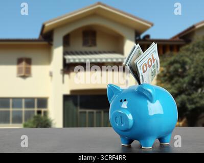 Sparen Sie Geld, um zu Hause zu kaufen. Blaue Sparkasse mit Dollars auf dem Tisch gegen Haus Stockfoto