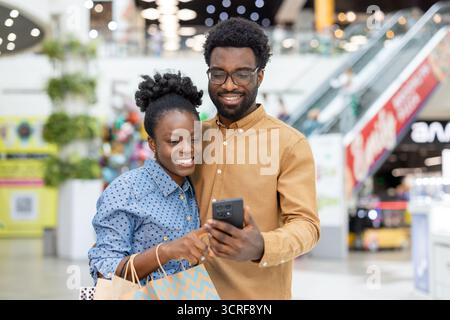 afroamerikanische Paare kaufen gemeinsam in einem geschäftigen Einkaufszentrum ein, lächeln und schauen sich ein Smartphone an und nutzen Technologie, um ihr Einkaufserlebnis zu verbessern Stockfoto