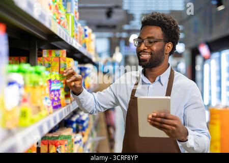 Junger schwarzer Mann auf einer Schürze, der in einem Supermarkt arbeitet, lächelt, während er Produkte in einem Regal überprüft und ein digitales Tablet für die Bestandsverwaltung hält Stockfoto