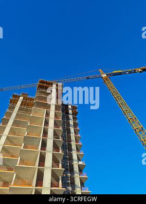 Weite Aufnahme eines gelben Turmkrains neben einem Betonhochhaus im Bau vor klarem, tiefblauem Himmel. Stockfoto