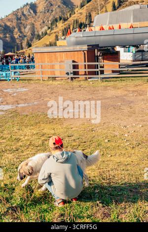 Eine junge kaukasische Frau mit blonden Haaren kniet auf Gras und spielt mit einem großen weißen Hund. Im Hintergrund sind Berge und ein Skilift zu sehen. Stockfoto