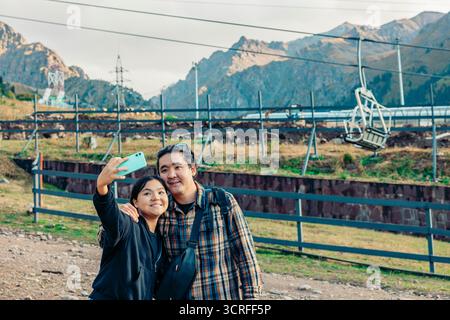 Eine junge Asiatin und ein asiatischer Mann mittleren Alters machen während ihres Urlaubs ein Selfie vor Bergen und einer Seilbahn. Stockfoto