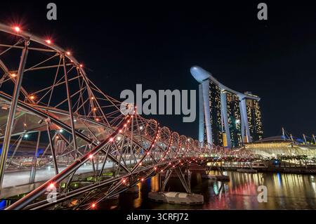 Blick auf die beleuchteten Stahlkurven der Helix Bridge, die in Richtung der berühmten Marina Bay Sands vor dem dunklen Nachthimmel führen, Singapur, Singapur. Stockfoto