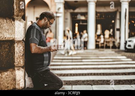Padua, italien 1. oktober 2025: Mann lehnt sich an eine Steinmauer auf dem belebten Bürgersteig der Stadt, fokussiert auf das Smartphone, während er in der Nähe eines Bürgersteigs wartete, ungezwungen urbanes Stockfoto