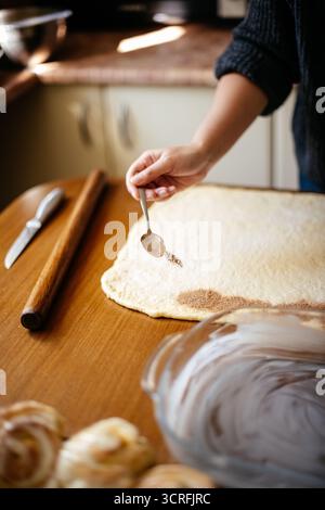 Eine Person, die Zimtzucker auf gerollten Teig streut, während sie in einer gemütlichen Küche hausgemachte Zimtrollen zubereitet Stockfoto