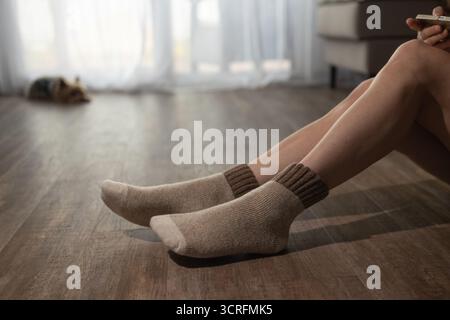 Eine Person sitzt auf einem Holzboden und trägt Socken. Ein Yorkshire Terrier liegt auf dem Boden im Hintergrund. Die Inneneinrichtung bietet Entspannung und Wärme. Stockfoto