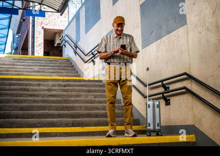 Glücklicher Seniorenmann steht am Bahnhof und sendet Nachrichten auf dem Smartphone. Senioren reisen. Stockfoto