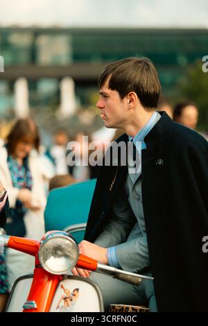London, Großbritannien – 2025.09.27 – Ein junger Mann in grauem Anzug und dunklem Mantel sitzt auf einem roten Oldtimer-Roller, der mit Retro-Kunstwerken im Classic Car B verziert ist Stockfoto