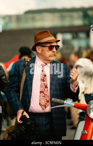 London, Großbritannien – 2025.09.27 – Ein Mann in kariertem Anzug, gemusterter Krawatte und braunem Fedora hält eine Kamera, während er bei einem Vintage-Roller im Classic steht Stockfoto