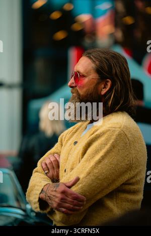 London, Großbritannien – 2025.09.27 – ein Mann mit langen Haaren, Vollbart, gelber Strickjacke und rosafarbener Sonnenbrille steht mit überkreuzten Armen am Classic Car Boot Stockfoto