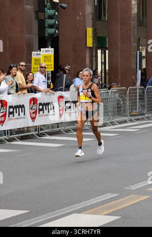 Marathonläuferin bewegt sich kraftvoll durch die Stadtstraße mit farbenfrohen Gebäuden, während begeisterte Zuschauer ihre Fitness feiern Stockfoto