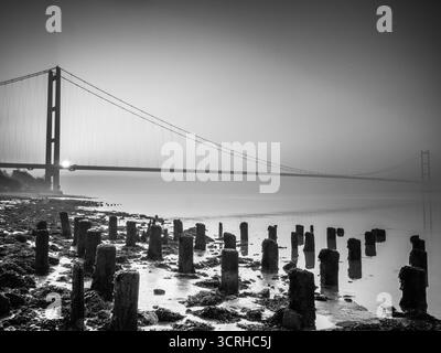 Die Humber Bridge erstreckt sich bei Sonnenaufgang in Schwarz-weiß über die Mündung mit alten Holzpfosten im Wattenmeer im Vordergrund Stockfoto