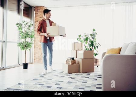 Junger Mann, der in eine moderne Wohnung zieht, mit Pappkartons in einem hellen Wohnzimmer voller Tageslicht Stockfoto