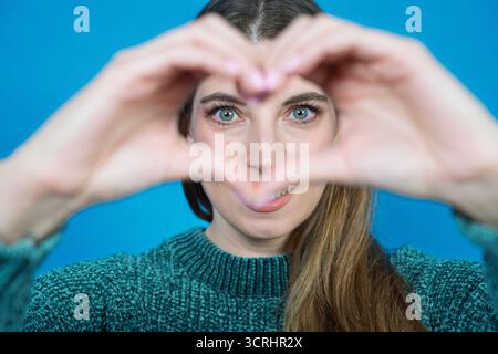 Frau, die mit Händen eine Herzgeste macht, zeigt Liebe und Zuneigung auf blauem Hintergrund Stockfoto