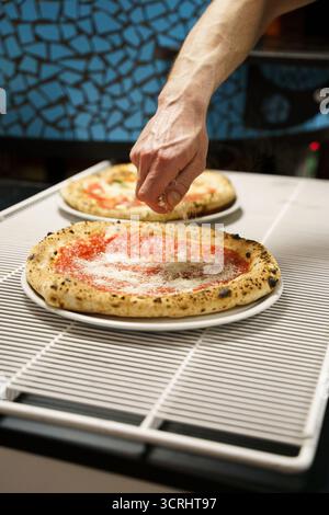 Nahaufnahme eines Küchenchefs, der geriebenen Käse über eine frisch gebackene Pizza im neapolitanischen Stil mit Tomatensoße streut. Zubereitung in einer authentischen Pizzeria Stockfoto