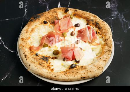 Blick von oben auf eine Gourmet-Pizza mit geschmolzenem Mozzarella, Schinkenscheiben und einer Mischung aus grünen und schwarzen Oliven. Stockfoto