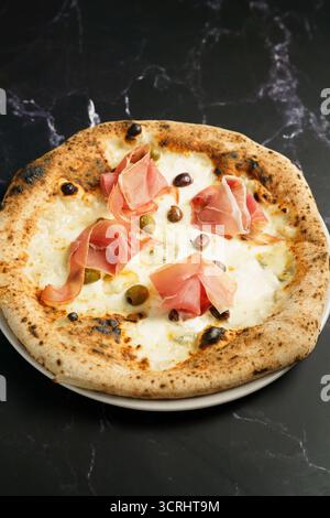 Blick von oben auf eine Gourmet-Pizza mit geschmolzenem Mozzarella, Schinkenscheiben und einer Mischung aus grünen und schwarzen Oliven. Stockfoto